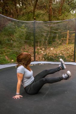 A young woman landing on her butt but safely on a trampoline in an outdoor setting.