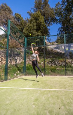 Mutlu bir şekilde gülümseyen genç, güçlü, atletik bir kadın, dağlarda oynanan açık havada oynanan tenis maçında topu yakalamak için zıplıyor..