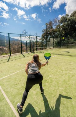 Güçlü genç bir sporcunun arka görüntüsü, çömelmiş ve raketiyle bir padel topuna vurmaya hazır açık bir dağ kortunun karşı tarafından ona yaklaşıyor..