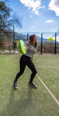 Genç, güçlü, mutlu bir atlet, gülümsüyor, gelen bir padel topuna vurmak üzere, raketi açık bir sahada, arka planda dağlık bir manzara ile..