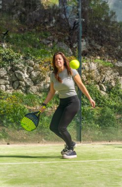 Bir padel topu, açık yeşil bir padel kortunda topu almaya hazır genç, atletik bir kadına doğru hızla yaklaşıyor..