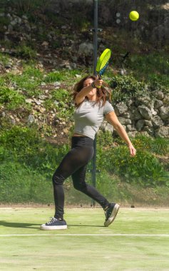 Genç, atletik bir kadın, açık havada bir padel kortunda forehand kullanarak, şık bir pozla bir padel topunu güçlü bir şekilde geri getiriyor..