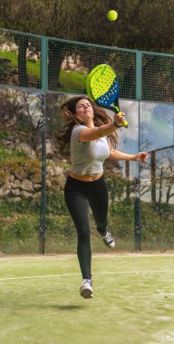 Padel oynayan genç ve güçlü bir atlet, topa raketle atlamak için zıplıyor ve açık yeşil bir kortta çok odaklanmış ve enerjik görünüyor..