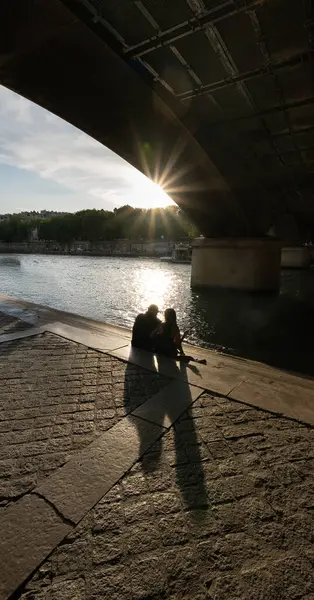 Paris 'te bir köprünün altında güneşle aydınlatılan romantik bir günbatımında Seine Nehri' nin kenarında oturan sevgi dolu bir çiftin arka plandaki silueti..