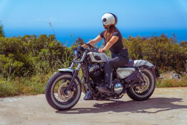 Güneşli bir günde okyanusa bakan manzaralı bir sahil yolunda Harley Davidson motosikleti süren bir kadın..