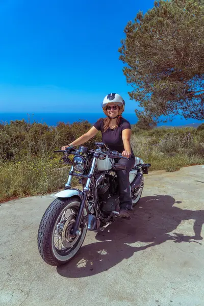 Güneşli bir günde arka planda deniz varken kıyı yolunda motosiklet sürerken mutlu bir şekilde gülümseyen genç kadın..