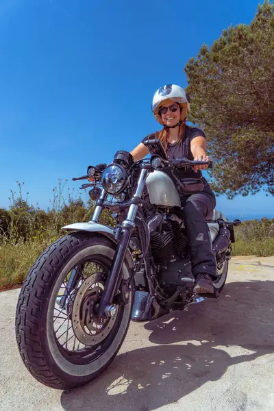 Güneşli bir günde mavi bir gökyüzünün altında kıyı yolu boyunca motosiklet sürerken kameraya gülümseyen kendine güvenen bir kadının geniş açılı görüntüsü..