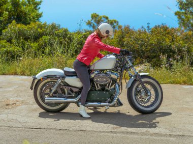 Kırmızı deri ceketli bir kadın Harley Davidson spor motosikletine biniyor güneşli bir sahil yolunda gezmeye hazırlanıyor..