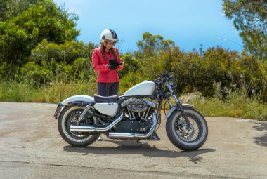 Kırmızı ceketli ve açık yüzlü bir kask takan güzel genç bir kadın özel Harley Davidson motosikletinin yanına eldivenlerini takıyor manzaralı bir sahil yolunda.