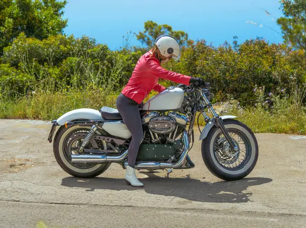 Kırmızı deri ceketli bir kadın Harley Davidson spor motosikletine biniyor güneşli bir sahil yolunda gezmeye hazırlanıyor..
