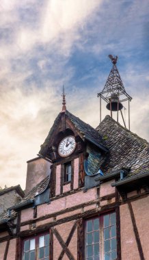 Tarihi bir saat kulesi ve bir horoz rüzgar gülü süslü bir demir çan kulesinin tepesinde Fransa 'nın Conques köyündeki yarı keresteli geleneksel bir evde, gün batımında mavi bir gökyüzünün altında..