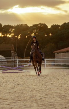 Genç bir İspanyol kadın binici, gün batımında kumlu bir tarlada atını engelli bir çitten atladıktan sonra mutlu ve mutlu bir şekilde gülümsüyor..