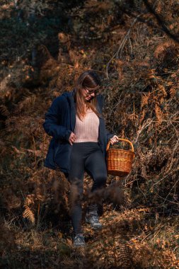 Young woman forager smiling enjoying happily walking downhill with a wicker woven basket through dense autumn foliage looking for mushrooms.