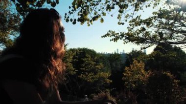 Uzak şehir siluetine bakan bir kadın. Ormanlık bir alanda oturuyor ve güneş yüzünde parlıyor..