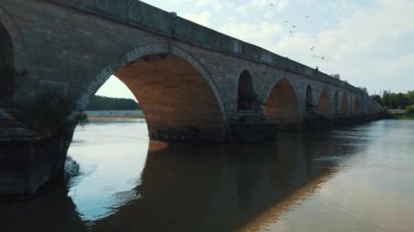 Osmanlı İmparatorluğu zamanından kalma taş bir köprü. Tunca Köprüsü. Kamera köprünün ayaklarına doğru uçar. Köprüden geçen insanlar ve uçan kuşlar görülür..