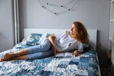 Genç, beyaz, güzel bir kadın kitap okuyor ya da koltukta ya da koltukta rahat rahat uzanıyor, mutlu ve rahat görünüyor. Gülümsüyor ve pencereden dışarı bakıyor. Yüksek kalite fotoğraf