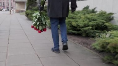 A man with a bouquet of red roses quickly goes to a meeting. Against the background of green bushes and buildings. Valentines Day. High quality 4k footage