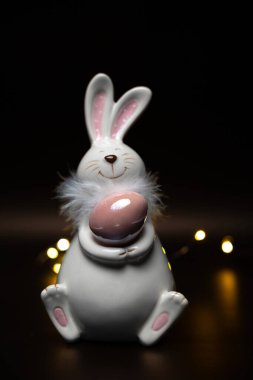 Easter white bunny with a brown egg on a black background with bright lights of garlands. Easter preparation. High quality photo