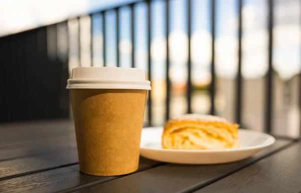 A paper cup of coffee with a white lid is on the table next to the pie. Cafe open veranda. High quality photo