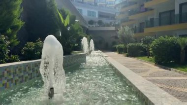 Konut binasının yanındaki çeşme, güneş altında parlayan su jetleri, manzara perspektifi. Yüksek kalite 4k görüntü