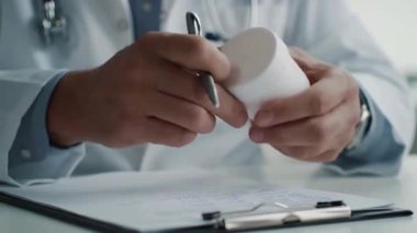 Doktor elinde kalemle masanın üstünde ilaç şişesi tutuyor.