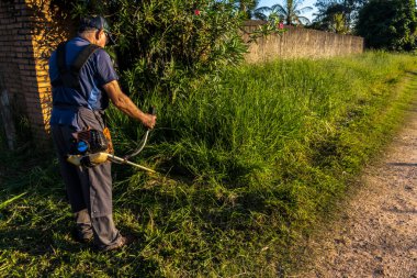 Marilia, Sao Paulo, Brezilya, 22 Mart 2023. İşçi, manuel benzin fırça kesicisiyle Marilia 'nın kırsal kesimindeki bir çiftliğin kapısındaki yağmurla birlikte büyüyen otları kesiyor.