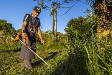 Marilia, Sao Paulo, Brezilya, 22 Mart 2023. İşçi, manuel benzin fırça kesicisiyle Marilia 'nın kırsal kesimindeki bir çiftliğin kapısındaki yağmurla birlikte büyüyen otları kesiyor.