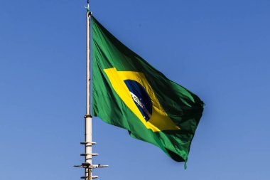 Arka planda mavi gökyüzü olan bir bayrak direğinde dalgalanan Brezilya bayrağı
