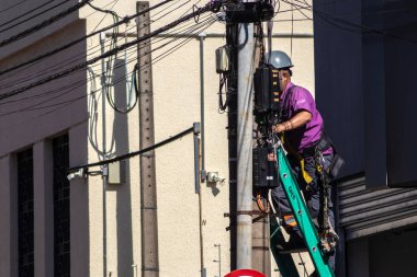 Marilia, Sao Paulo, Brezilya, 12 Haziran 2024. Bir telefon, kablo TV ve internet şirketi çalışanı, Marilia şehrindeki bir direğe kablo döşeyen bir merdivende çalışıyor.