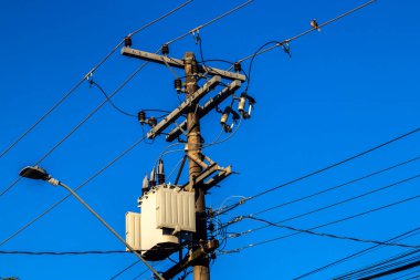 Brezilya 'da transformatörlü elektrik direği ve açık mavi gökyüzünün altındaki kablolar. Kentsel güç dağılımı altyapısını gösteren bu resim.