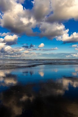 Sakin bir sahil sahnesi. Bulutlar, derin mavi gökyüzünün altındaki ıslak kuma mükemmel bir şekilde yansıyor. Huzurlu ve simetrik bir doğal manzara yaratıyor.