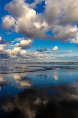 Sakin bir sahil sahnesi. Bulutlar, derin mavi gökyüzünün altındaki ıslak kuma mükemmel bir şekilde yansıyor. Huzurlu ve simetrik bir doğal manzara yaratıyor.