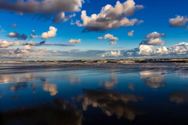 Sakin bir sahil sahnesi. Bulutlar, derin mavi gökyüzünün altındaki ıslak kuma mükemmel bir şekilde yansıyor. Huzurlu ve simetrik bir doğal manzara yaratıyor.