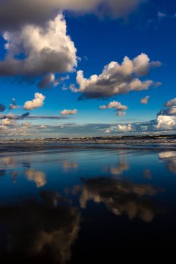 Sakin bir sahil sahnesi. Bulutlar, derin mavi gökyüzünün altındaki ıslak kuma mükemmel bir şekilde yansıyor. Huzurlu ve simetrik bir doğal manzara yaratıyor.