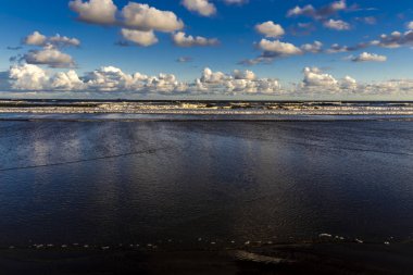 Sakin bir sahil sahnesi. Bulutlar, derin mavi gökyüzünün altındaki ıslak kuma mükemmel bir şekilde yansıyor. Huzurlu ve simetrik bir doğal manzara yaratıyor.