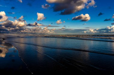 Sakin bir sahil sahnesi. Bulutlar, derin mavi gökyüzünün altındaki ıslak kuma mükemmel bir şekilde yansıyor. Huzurlu ve simetrik bir doğal manzara yaratıyor.