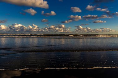Sakin bir sahil sahnesi. Bulutlar, derin mavi gökyüzünün altındaki ıslak kuma mükemmel bir şekilde yansıyor. Huzurlu ve simetrik bir doğal manzara yaratıyor.