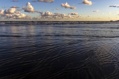 Sakin bir sahil sahnesi. Bulutlar, derin mavi gökyüzünün altındaki ıslak kuma mükemmel bir şekilde yansıyor. Huzurlu ve simetrik bir doğal manzara yaratıyor.
