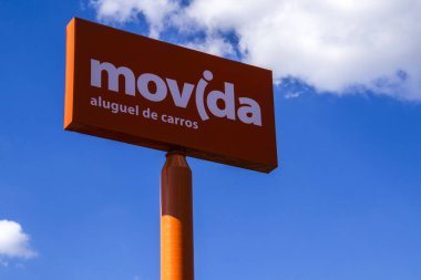 Marilia, Sao Paulo, Brezilya. 12 Mart 2025. Mavi gökyüzünün altında beyaz bulutlu bir araba kiralama tabelası. Brezilya 'da şirket logosu olan parlak turuncu tabela. Portekizce bir araba kiralayın.