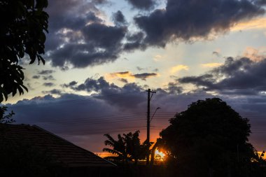 Brezilya 'nın kırsal kesiminde canlı bir günbatımı. Ağaçların siluetleri ve sıcak gökyüzü renkleriyle mimari kontrastlar