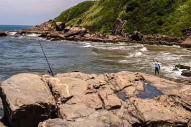 Itanhaem, Sao Paulo, Brezilya. 22 Ocak 2018 'de kayalık sahilde bir balıkçı, balık tutuyor ve okyanusu seyrediyor. İnsanoğlu ve doğa arasındaki bağı yakalar, kıyı manzaraları ve deniz sükuneti