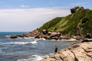Itanhaem, Sao Paulo, Brezilya. 22 Ocak 2018 'de kayalık sahilde bir balıkçı, balık tutuyor ve okyanusu seyrediyor. İnsanoğlu ve doğa arasındaki bağı yakalar, kıyı manzaraları ve deniz sükuneti