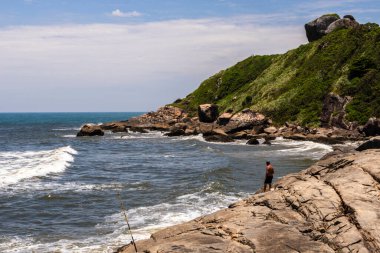 Itanhaem, Sao Paulo, Brezilya. 22 Ocak 2018 'de kayalık sahilde bir balıkçı, balık tutuyor ve okyanusu seyrediyor. İnsanoğlu ve doğa arasındaki bağı yakalar, kıyı manzaraları ve deniz sükuneti