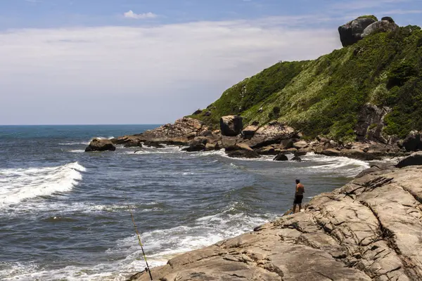 Itanhaem, Sao Paulo, Brezilya. 22 Ocak 2018 'de kayalık sahilde bir balıkçı, balık tutuyor ve okyanusu seyrediyor. İnsanoğlu ve doğa arasındaki bağı yakalar, kıyı manzaraları ve deniz sükuneti