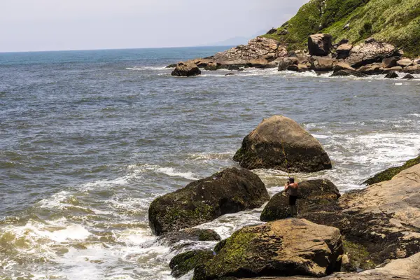 Itanhaem, Sao Paulo, Brezilya. 22 Ocak 2018 'de kayalık sahilde bir balıkçı, balık tutuyor ve okyanusu seyrediyor. İnsanoğlu ve doğa arasındaki bağı yakalar, kıyı manzaraları ve deniz sükuneti
