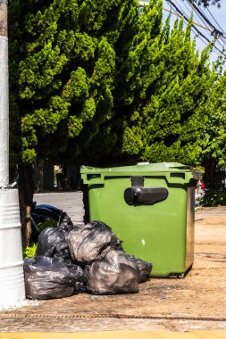 Yeşil çöp konteynırları ve siyah çöp torbaları yapraklı bir sokak kaldırımında. Atık yönetimi, kentsel hijyen ve çevresel farkındalık.