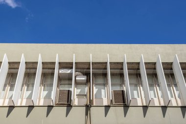 Dikey güneş gözlüklü, pencere ızgaralı ve mavi gökyüzüne karşı klimalı modernist bina cephesi.