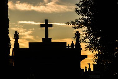 Mezarlık siluetleri alacakaranlıkta, renkli gökyüzüne karşı haçlar ve heykellerle. İnanç, hafıza ve yansıma temalarını çağrıştırır