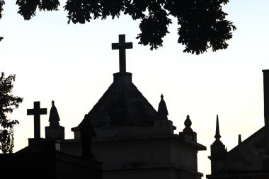 Mezarlık siluetleri alacakaranlıkta, renkli gökyüzüne karşı haçlar ve heykellerle. İnanç, hafıza ve yansıma temalarını çağrıştırır