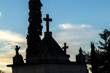 Mezarlık siluetleri alacakaranlıkta, renkli gökyüzüne karşı haçlar ve heykellerle. İnanç, hafıza ve yansıma temalarını çağrıştırır